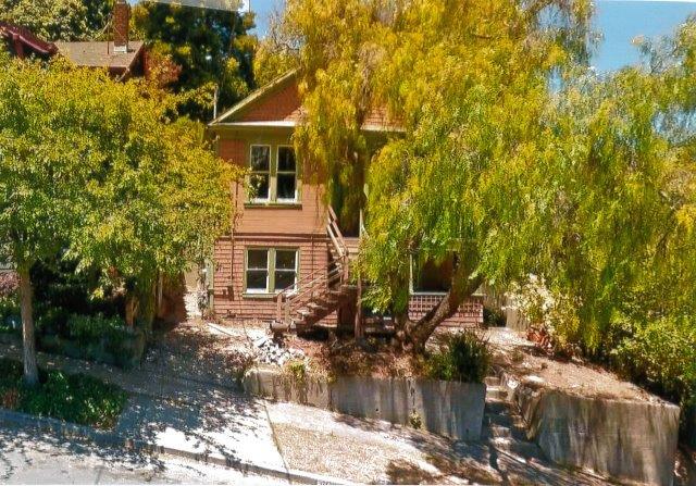 Before: Front view of house across the street steeply sloped down to the right. Crumbling retaining wall and obscuring trees are visible.